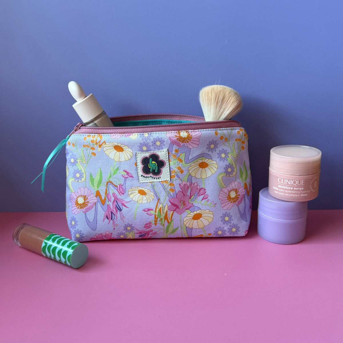 Wash bag with a pastel retro inspired floral print, ByLouMoffat floral logo labels in the top middle of the bag and rose coloured zip with a teal ribbon pull. Displayed on a pink surface against a lilac background surrounded by cosmetic items including a pink and purple moisturiser jar, and a lip gloss tube.