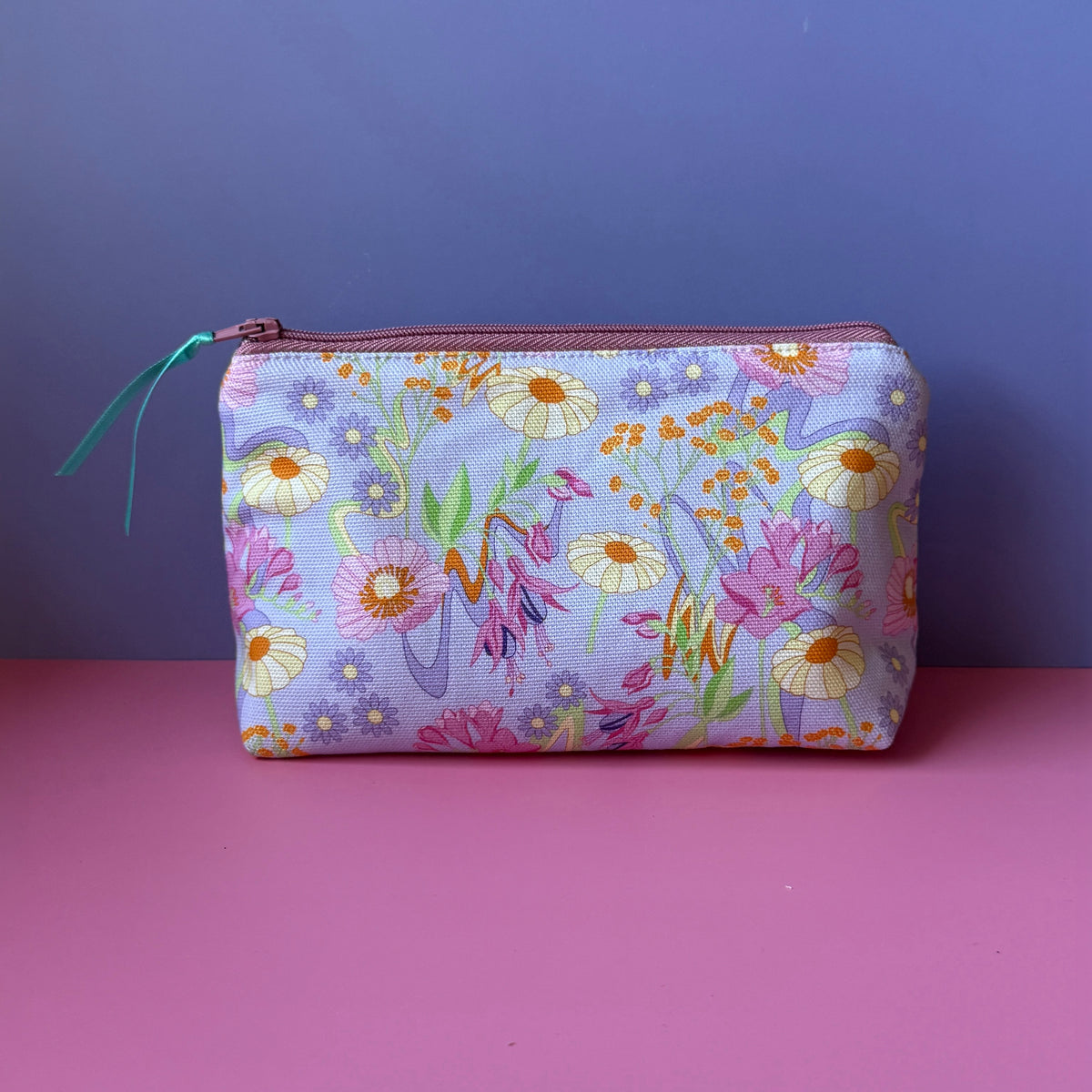 A wash bag with a pastel retro inspired floral print and rose coloured zip with a teal ribbon pull. Displayed on a pink surface against a lilac background.