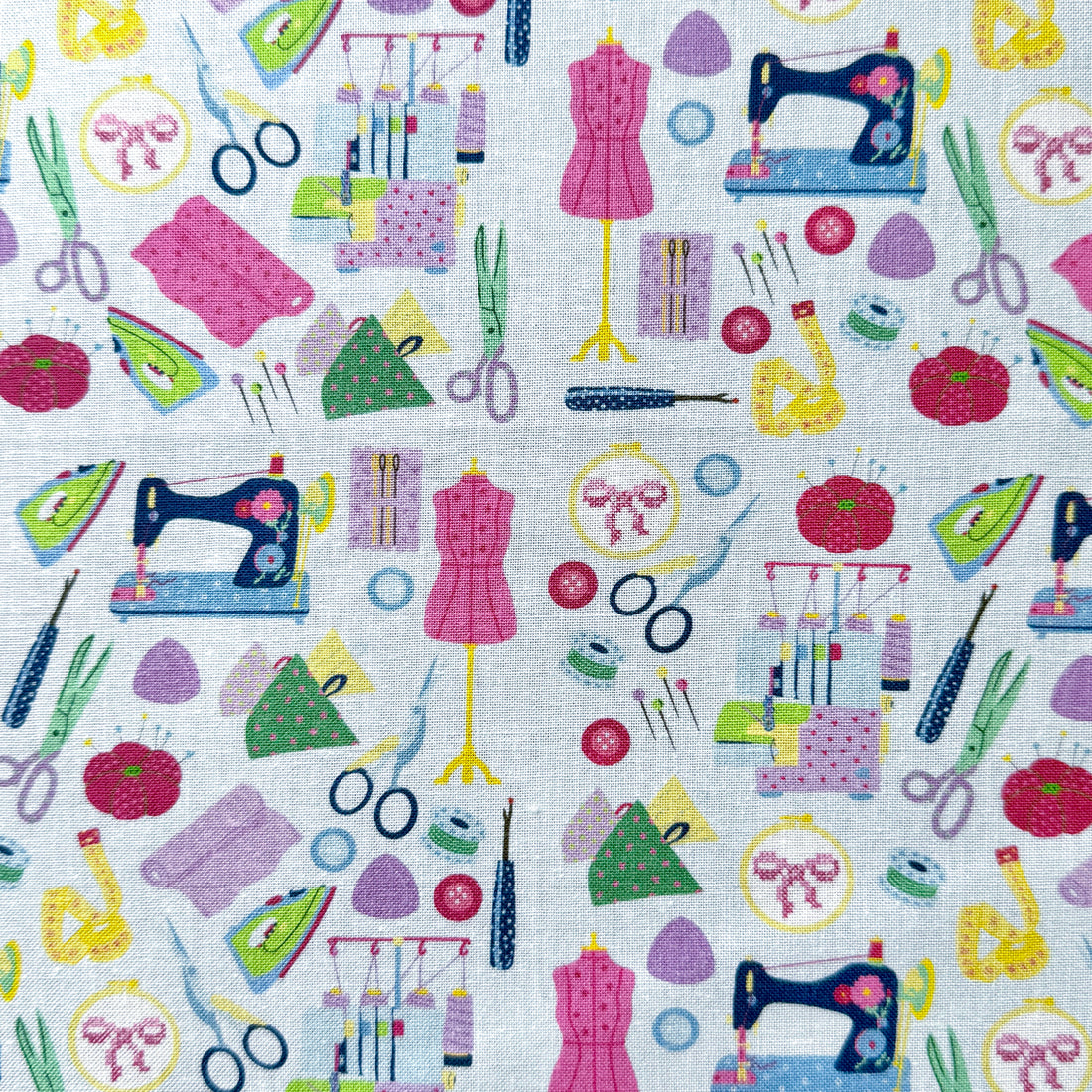 Sewing-themed fabric laid flat, featuring colourful sewing machines, scissors, dress forms, pin cushions, tape measures and irons on a light blue background.