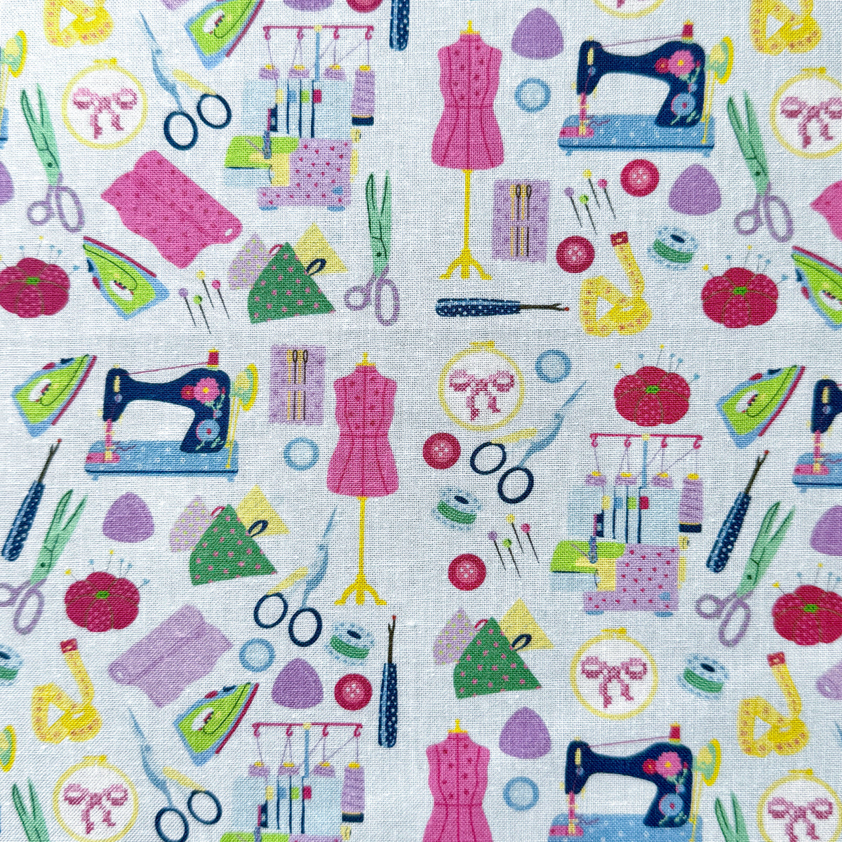 Sewing-themed fabric laid flat, featuring colourful sewing machines, scissors, dress forms, pin cushions, tape measures and irons on a light blue background.