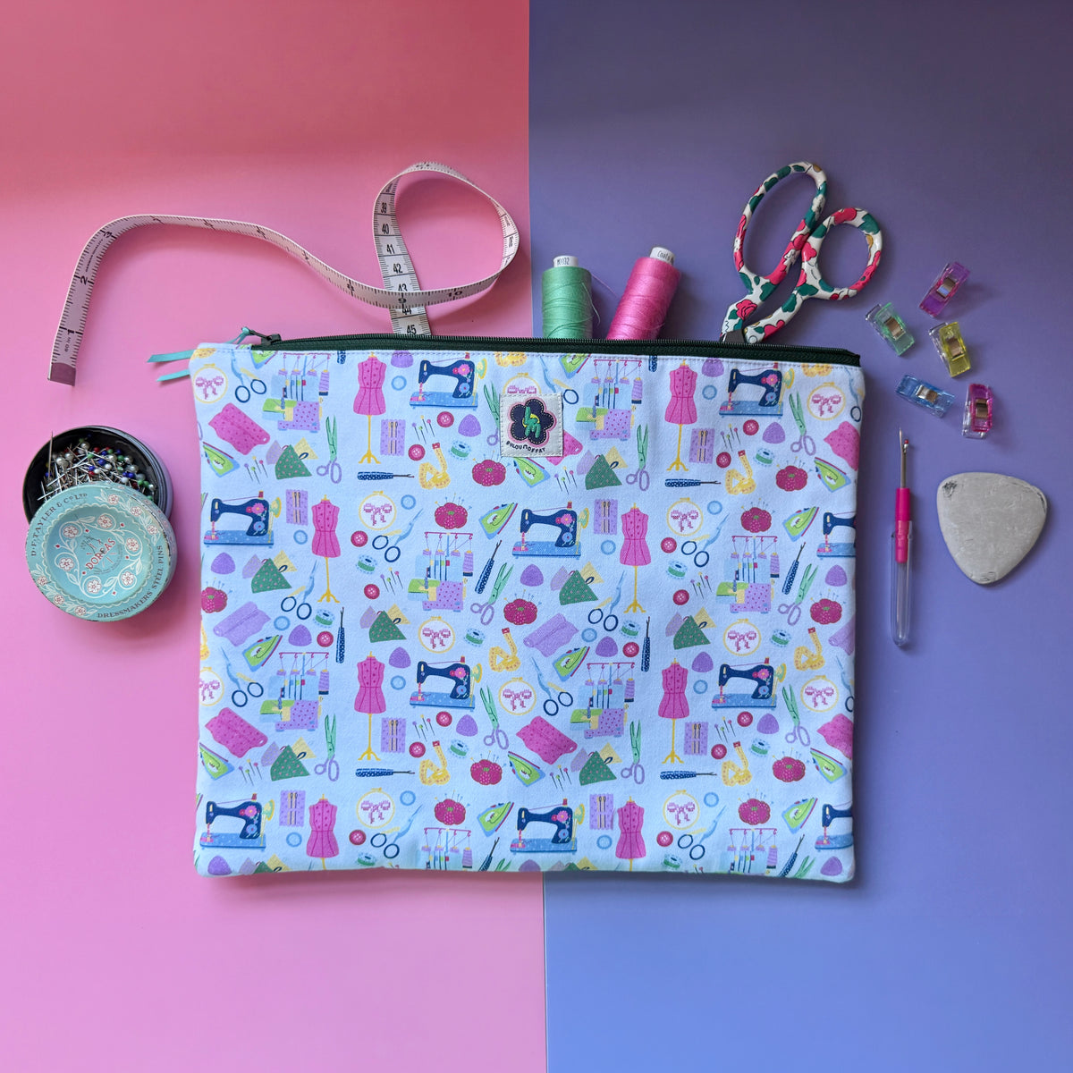 Large sewing-print zip pouch displayed with a tape measure, scissors, thread, pins and sewing clips. Fabric features colourful sewing machines, scissors, dressmaking mannequins, embroidery hoops, unpickers and other sewing notions on a pale blue background. 