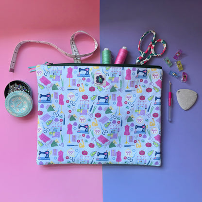 Large sewing-print zip pouch displayed with a tape measure, scissors, thread, pins and sewing clips. Fabric features colourful sewing machines, scissors, dressmaking mannequins, embroidery hoops, unpickers and other sewing notions on a pale blue background. 