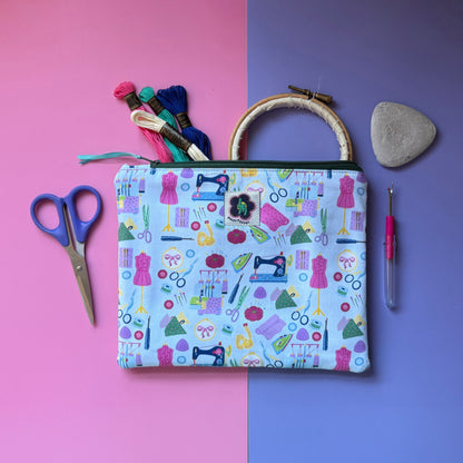 Medium sewing-print zip pouch displayed with embroidery threads, an embroidery hoop, scissors and tailor’s chalk. Fabric features colourful sewing machines, scissors, dressmaking mannequins, embroidery hoops, unpickers and other sewing notions on a pale blue background. 