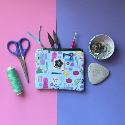 Small pale blue sewing-print pouch with a green zip, displayed with scissors, thread, pins and tailor’s chalk. Fabric features colourful sewing machines, scissors, dressmaking mannequins, embroidery hoops, unpickers and other sewing notions on a pale blue background. 
