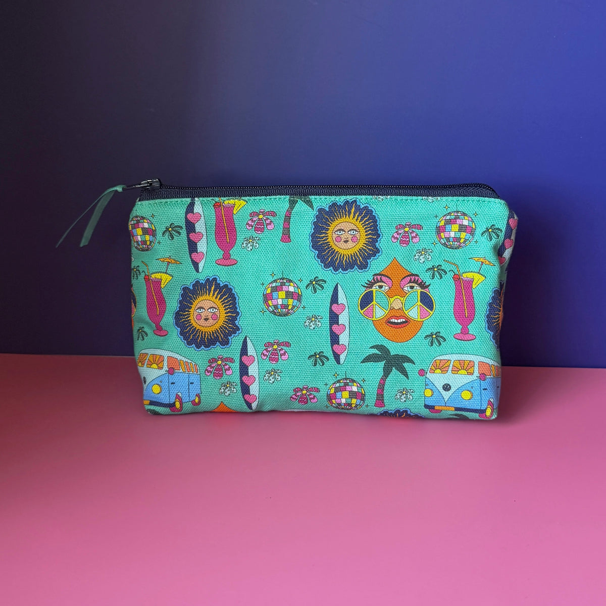 A hippie makeup bag with a a colourful  hippie inspired design featuring camper vans, surf boards, cocktails, sun, palm tress and disco ball. It has a navy blue zip with teal ribbon sits on a pink surface against a royal blue background. 