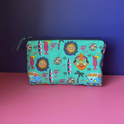 A hippie makeup bag with a a colourful  hippie inspired design featuring camper vans, surf boards, cocktails, sun, palm tress and disco ball. It has a navy blue zip with teal ribbon sits on a pink surface against a royal blue background. 