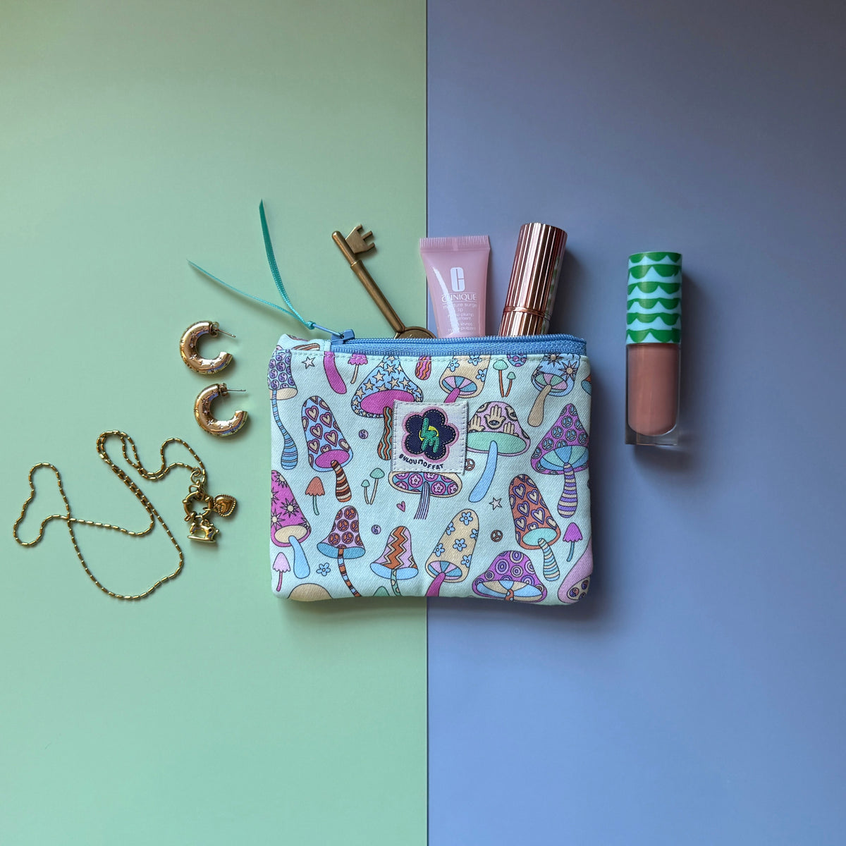 Small mushroom-print zip pouch shown with cosmetics, a key, gold hoop earrings and a necklace. The design features colourful patterned mushrooms featuring stars, hearts, flowers hamsa hands. 