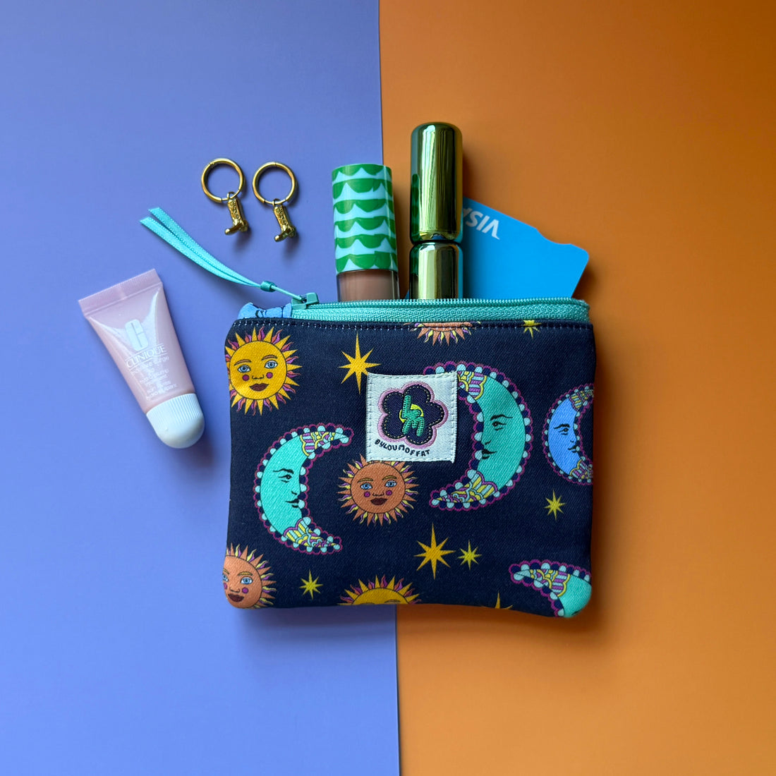 Small navy sun and moon print pouch with a turquoise zip, shown with cosmetics, earrings and a bank card. The fabric the pouch is made from features a navy background with teal/blue moons, orange suns and yellow stars. 