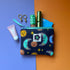 Small navy sun and moon print pouch with a turquoise zip, shown with cosmetics, earrings and a bank card. The fabric the pouch is made from features a navy background with teal/blue moons, orange suns and yellow stars. 