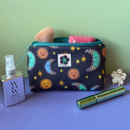 A celestial makeup bag with a navy and teal design that features suns, moons and stars sits open on a lilac surface against a light green background, surrounded by cosmetic items including a green mascara tube, a small floral hair clip and a small mirrored spray bottle. 