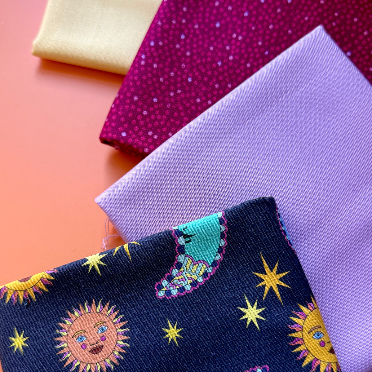 Four folded fat quarters arranged in a square, featuring a navy celestial print, pale yellow, lilac and berry pink fabrics.