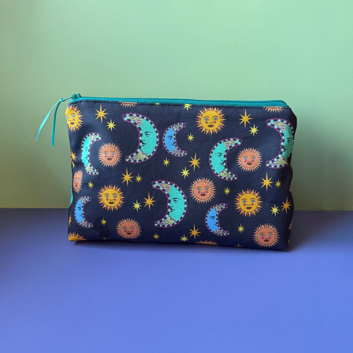 A celestial makeup bag with a navy, orange, blue and teal design that features suns, moons and stars,  sits on a lilac surface with light green backdrop. The pouch features a pattern of suns, moons and stars with a navy base, with a  teal zip.