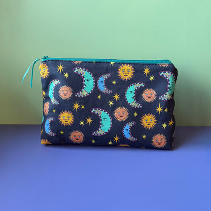 A celestial makeup bag with a navy, orange, blue and teal design that features suns, moons and stars,  sits on a lilac surface with light green backdrop. The pouch features a pattern of suns, moons and stars with a navy base, with a  teal zip.