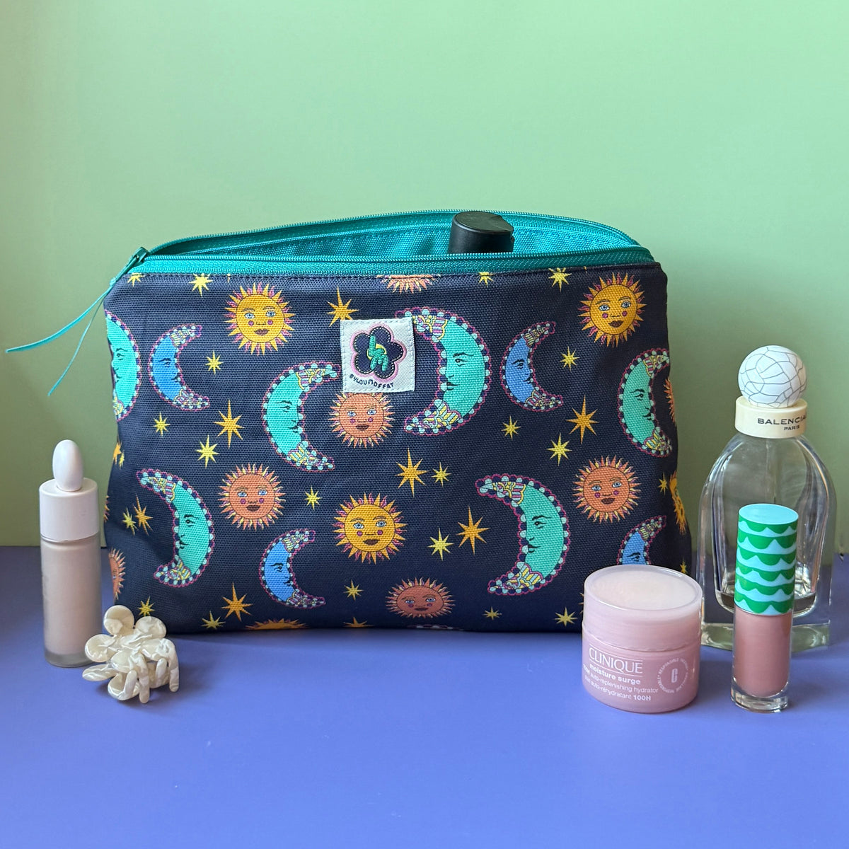 A  zippered wash bag/makeup bag with a navy and teal celestial design featuring suns, moons and stars sits on a lilac surface against a light green background, surrounded by small beauty accessories including a Balenciaga Paris perfume bottle, lip gloss tube, pink jar,  small white floral hair clip and concealer tube.
