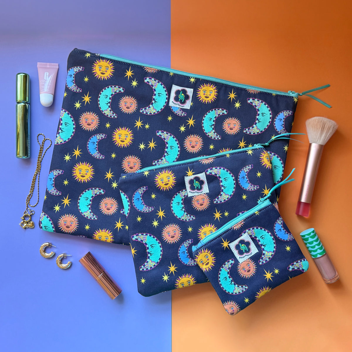 Three navy celestial print pouches with turquoise zips, surrounded by cosmetics, a makeup brush and jewellery.  The fabric the pouches is made from features a navy background with teal/blue moons, orange suns and yellow stars. 