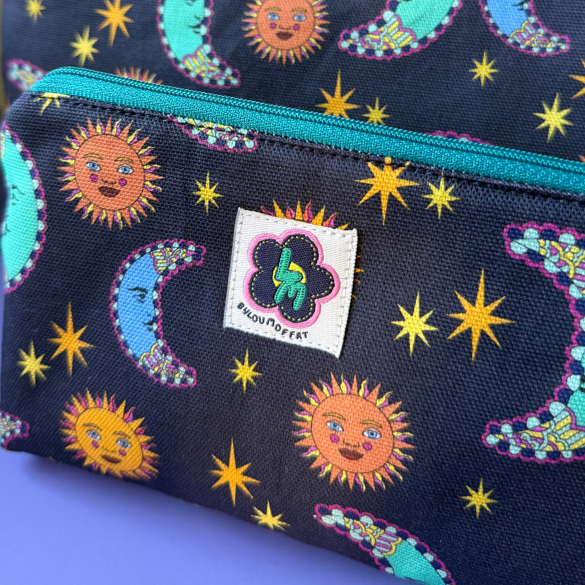 Close-up of a celestial zip pouch with two matching bags, featuring a sun and moon design on a navy background with a teal zipper and a visible ByLouMoffat brand label.