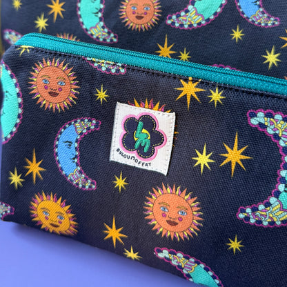 Close-up of a celestial zip pouch with two matching bags, featuring a sun and moon design on a navy background with a teal zipper and a visible ByLouMoffat brand label.