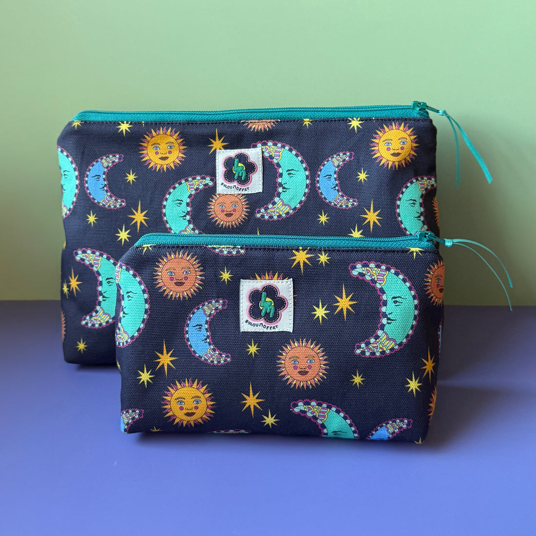 Two fabric zip pouches with a navy fabric featuring teal and blue moons, orange stars and yellow stars.  ByLouMoffat floral logo labels in the top centre of each bag and teal zip with a teal ribbon pull. Displayed on a lilac surface against a light green background.